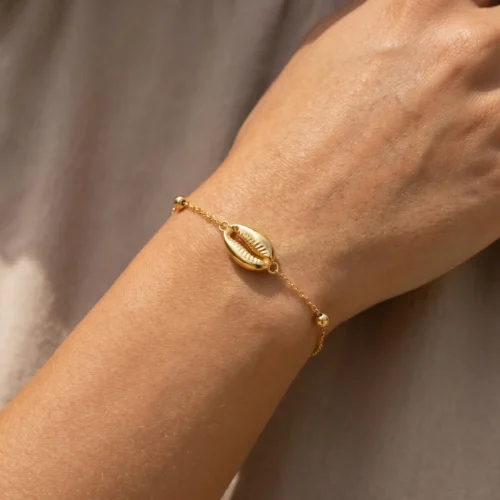 Craquez pour ce bracelet fin en Or massif 18K. Motif coquillage (Wda3) élégant, chaîne délicate et petite Khmisa porte-bonheur. Livraison au Maroc. Texte ALT image Bracelet fin en chaîne d'or massif 18 carats avec un motif central en forme de coquillage cauri, de petites perles dorées et un mini pendentif Khmisa près du fermoir, sur fond noir.