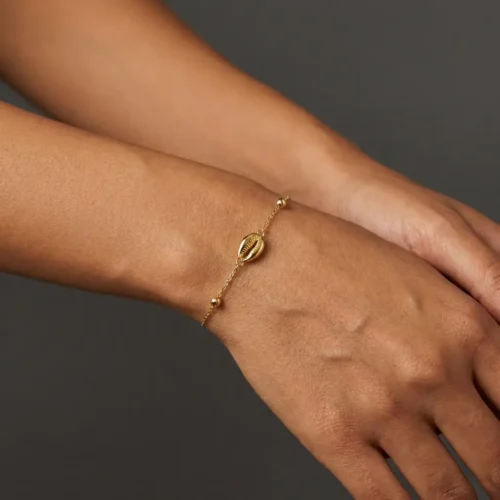 Craquez pour ce bracelet fin en Or massif 18K. Motif coquillage (Wda3) élégant, chaîne délicate et petite Khmisa porte-bonheur. Livraison au Maroc. Texte ALT image Bracelet fin en chaîne d'or massif 18 carats avec un motif central en forme de coquillage cauri, de petites perles dorées et un mini pendentif Khmisa près du fermoir, sur fond noir.