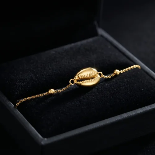 Craquez pour ce bracelet fin en Or massif 18K. Motif coquillage (Wda3) élégant, chaîne délicate et petite Khmisa porte-bonheur. Livraison au Maroc. Texte ALT image Bracelet fin en chaîne d'or massif 18 carats avec un motif central en forme de coquillage cauri, de petites perles dorées et un mini pendentif Khmisa près du fermoir, sur fond noir.
