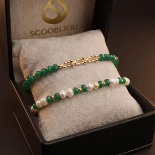 Duo de bracelets ajustables en macramé vert foncé ornés de véritables perles d'eau douce, de pierres vertes facettées et de finitions géométriques en or 18K, présentés dans un écrin noir.